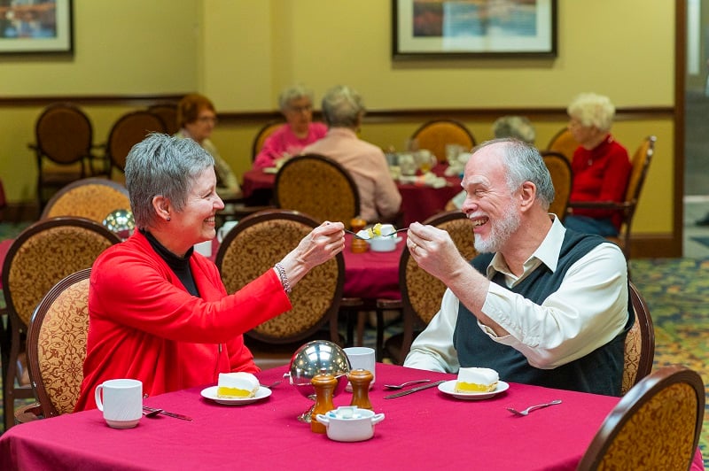 Senior woman in red jacket and man in vest eat lemon meringue pie together at table