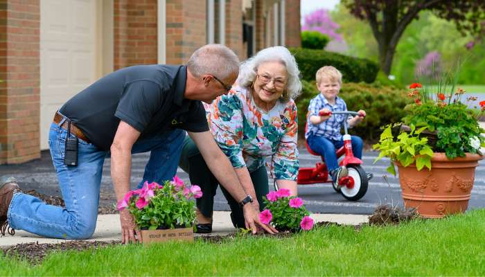 Senior couple plants flowers outside while grandson rides trike nearby