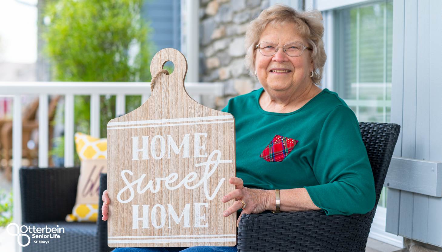 Otterbein St. Marys resident sitting on her front porch holding a sign that says “home sweet home.”